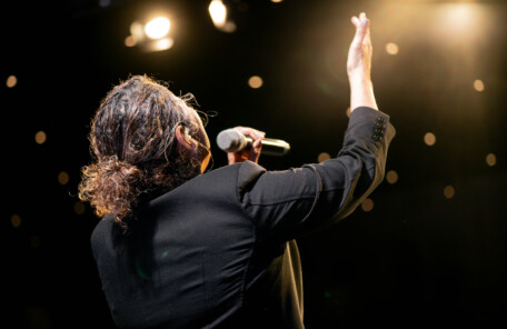 ライブステージでマイクを握り手を挙げて歌うボーカリストの背面ショット