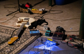 床に置かれたエレキギターと複数のエフェクターペダル、配線されたケーブルが写る演奏準備中のセッティング