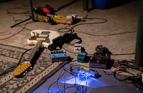 床に置かれたエレキギターと複数のエフェクターペダル、配線されたケーブルが写る演奏準備中のセッティング