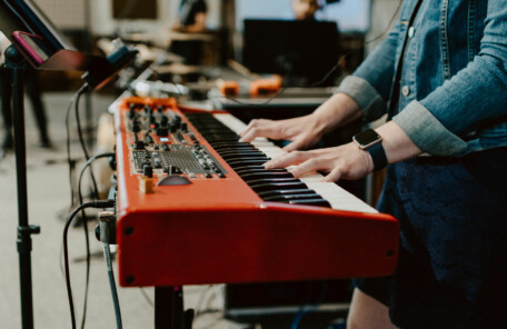 スタジオ内で赤いキーボードを両手で演奏している様子と、周囲に設置された機材が写っている