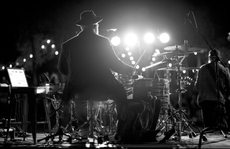 ステージ上で照明を背に、ドラムセットを演奏している演奏者の後ろ姿と周囲の機材が写ったモノクロ写真