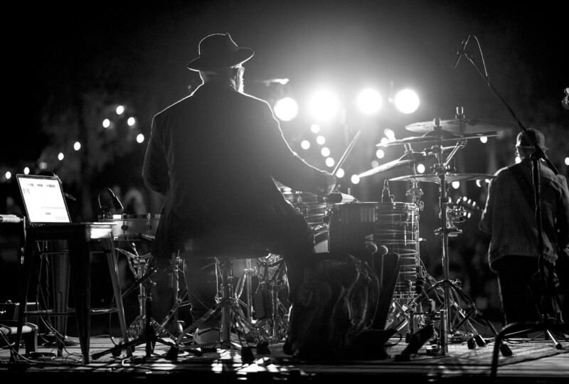 ステージ上で照明を背に、ドラムセットを演奏している演奏者の後ろ姿と周囲の機材が写ったモノクロ写真